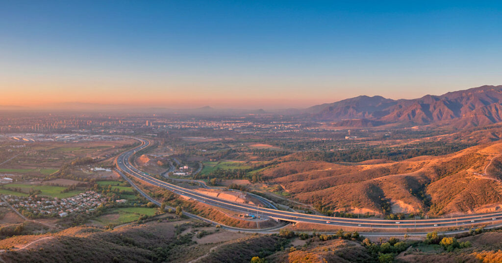 Vista panorámica simulada de una autopista atravesando un paisaje montañoso y urbano en Maipú, Chile, durante el atardecer.